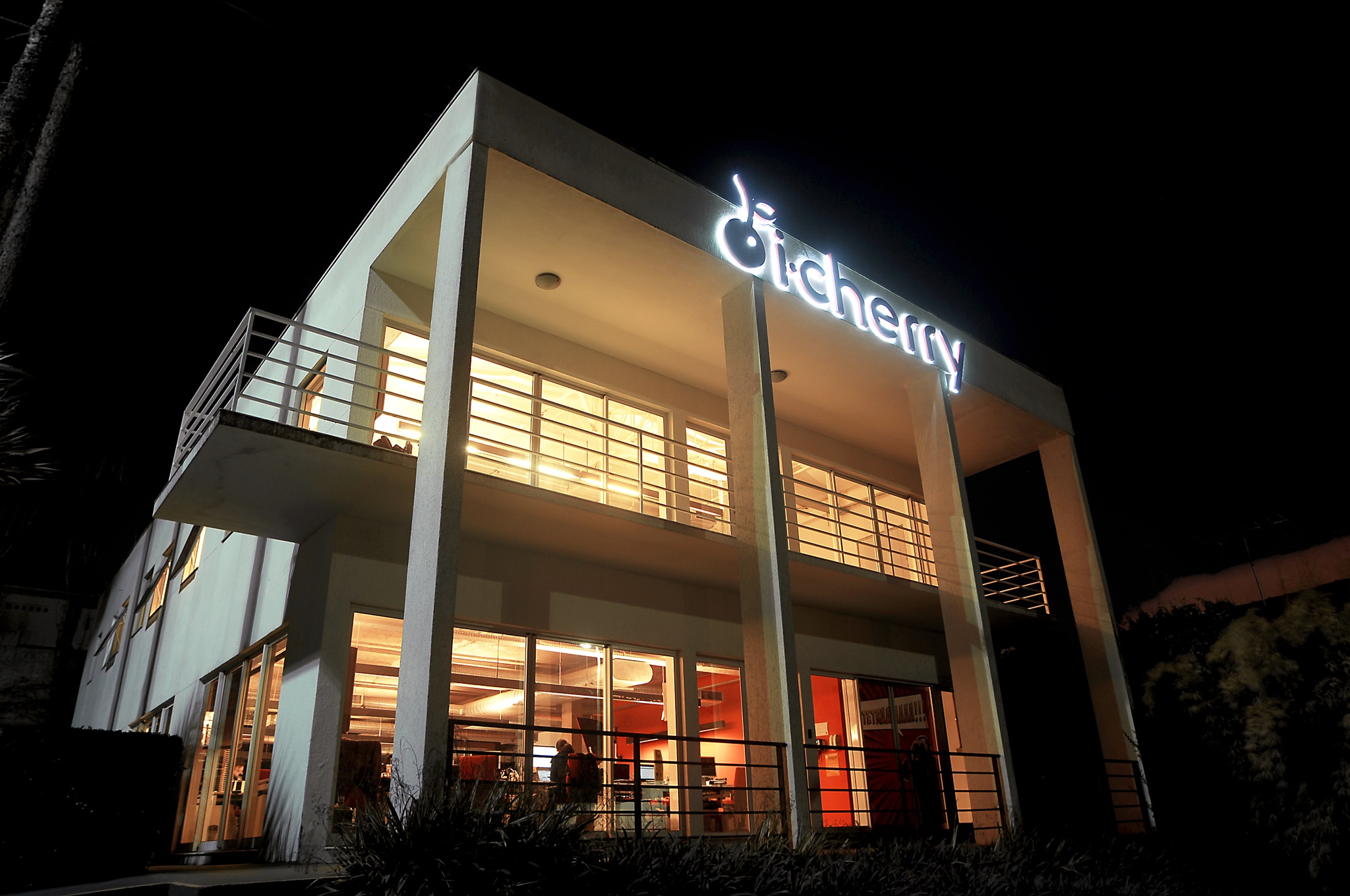 Foto da agencia da i-Cherry, um prédio branco com salas amplas grandes portas de vidro e uma grande logo da empresa luminosa na fachada
